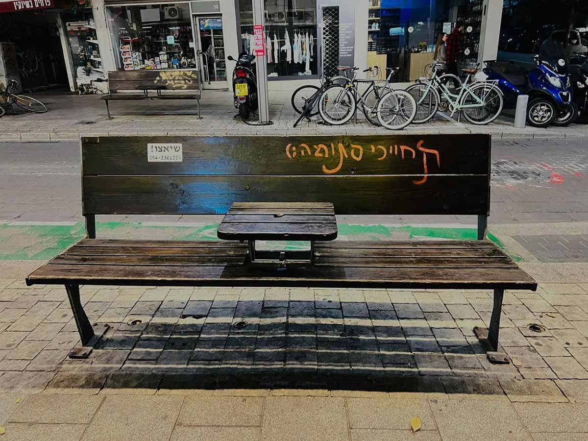 גרפיטי על ספסל רחוב הצבוע בחום, ועל משענת הגב של הספסל כתובת בצבע כתום: תחייכי סתומה : ). בצד שמאל של המשענת שלט פרסומת של שיאצו ! עם מספר טלפון. ברקע המדרכה בצד השני של הכביש עם חניית אופניים ואופנועים בצד ימין, וספסל דומה בצד שמאל. וברקע חנויות