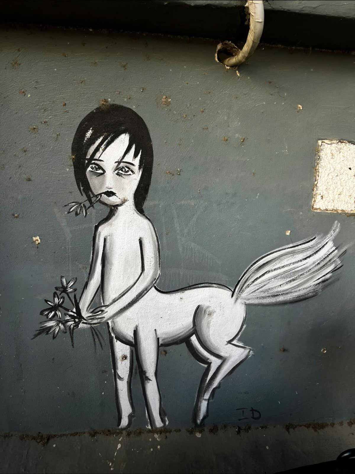 גרפיטי של סוס אישה, סוס עם זנב גדול, וכלג גוף עליון של אישה עם פרח בפה וזר פרחים בידיים. ציור בלבן ושחור על רקע של מלבן אפור.