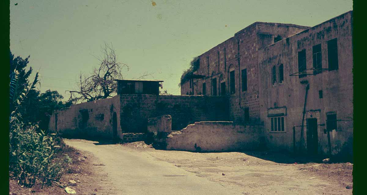 בית השייח' של שייח' מוניס בשנת 1964, צילום בשחור לבן. בניין בן שלוש קומות בצד ימין ובחזיתו מבנה של קומה אחת. ליד הבניין רחוב לא סלול ובצד שמאל צמחייהץ