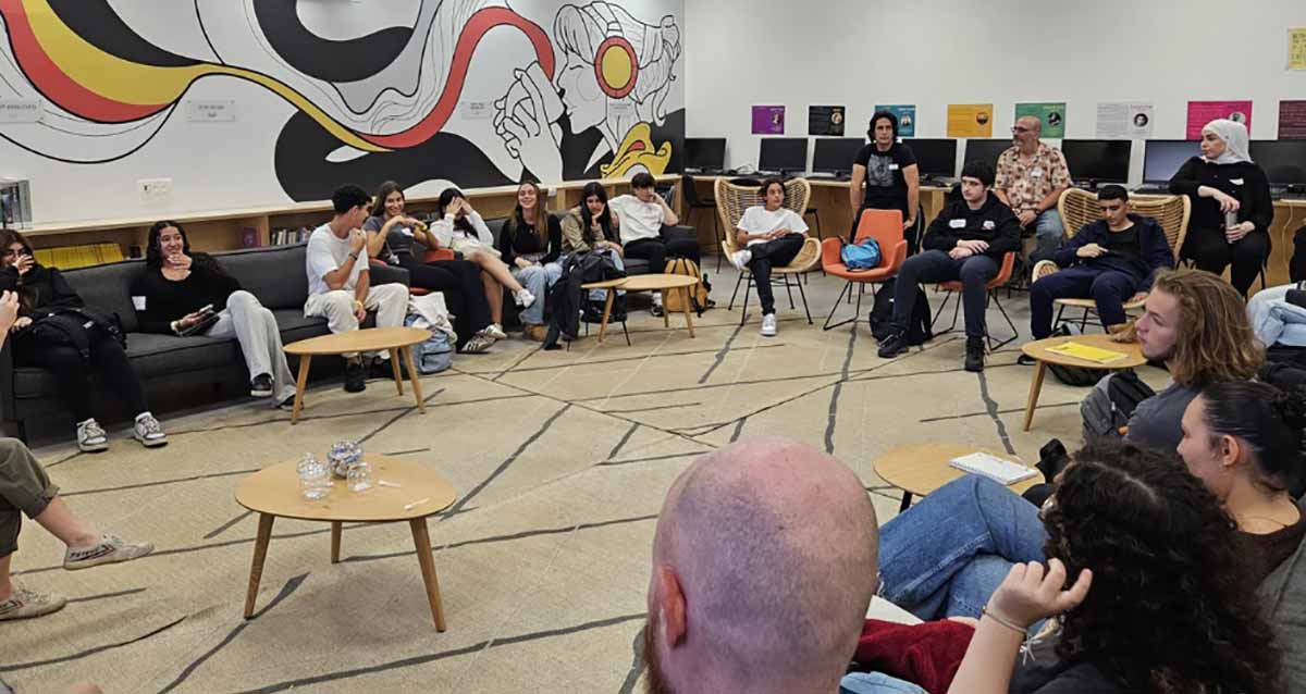 כנס תלמידים בתיכון שמיר. התלמידים יושבים במעגל עם מורים, בחדר גדול. בתוך המעגל שולחנות קטנים. על קיר אחד ציור על כולו, בצד ימין מצוירת בחורה שותה מכוס, ומהכוס יוצא פס אדום שהופך לצהוב ובהמשך מתפצל לארבעה צבעים: לבן, אדום, צהוב ושחור.