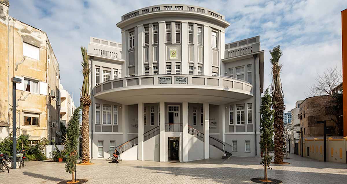 מוזיאון העיר תל אביב יפו בכיכר ביאליק. בניין של 4 קומות עם חזית מעוגלת. מדרגות אל הקומה השנייה בחזית מימין ומשמאל. עצי דקל מימין ומשמאל למוזיאון.