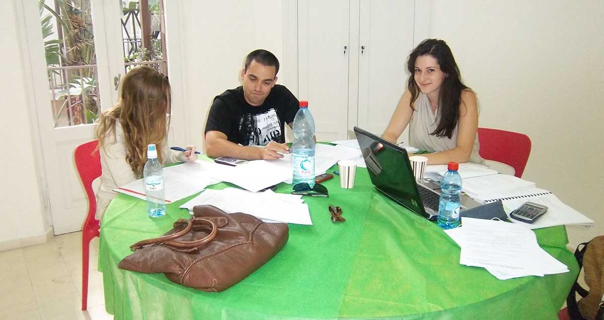 מרחב למידה לסטודנטים. שתי סטודנטיות וסטודנט אחד יושבים סביב שולחן עגול ועליו מפה ירוקה. על השולחן מחשב נייד, מחברות, ניירות, סלולרי, תיק חום ובקבוקי מים.