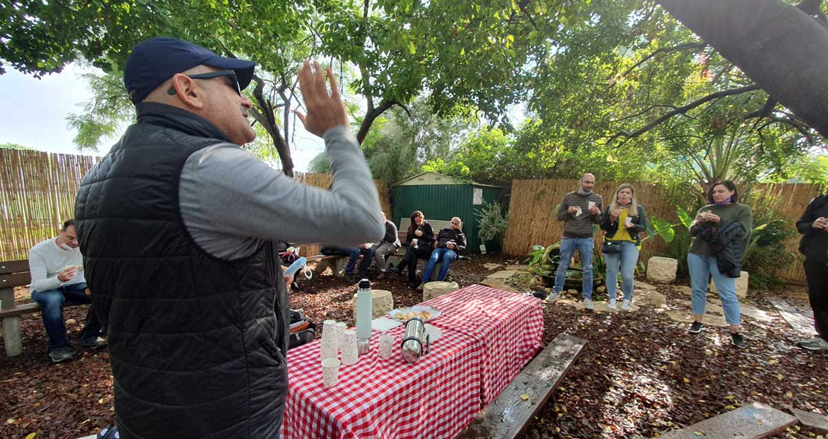 סיור קולינארי בתל אביב. המדריך עומד בגינה ליד שולחן עם כוסות מים, והמטיילים חלקם יושבים מולו על ספסלים וחלקם עומדים.