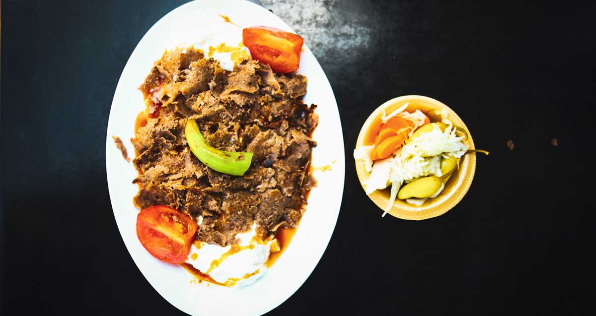 מנת איסקנדר בשווארמה מוטאפק. בצלחת בשר חתוך דק, חצאי עגבניות וקישוא. לידה צלחת קטנה עם חמוצים.