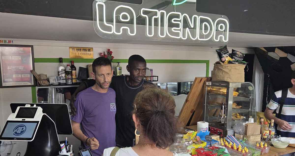 המכולת הלאטינית לה טיינדה בת"א. שני מוכרים וקונה אחת. מעל המוכרים שלט מואר באנגלית: LA TIENDA